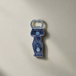 This metallic bottle opener features a Portuguese girl in a kneeling pose, her hands behind her head. Her bikini is adorned with the national colors and emblem of Portugal, and "Portugal" is written prominently at the top. A magnet on the back adds functionality for easy placement on metal surfaces.