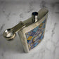 A stainless steel flask featuring a vibrant patchwork of blue, yellow, and white tile designs with "PORTUGAL" written boldly in the center. The cap is connected by a small hinge for easy use, while the flask rests against a white background, casting a gentle shadow on one side.
