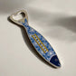A metal bottle opener shaped like a fish, featuring detailed blue and white patterns inspired by Portuguese azulejos tiles. The word "Portugal" is clearly visible on its side, and a small magnet on the reverse side allows it to attach to your fridge. The bottle opener function is located at the tail of the fish.