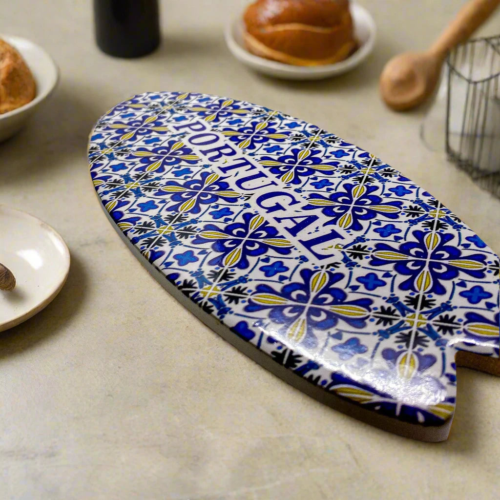 A beautifully crafted oval ceramic object adorned with a lively mosaic pattern of traditional azulejo tiles from Portugal. In the center, "PORTUGAL" is boldly displayed in blue uppercase letters. The souvenir has a cork base and casts a gentle shadow on a white surface.