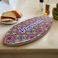 An oval ceramic souvenir decorated with a vibrant mosaic pattern reminiscent of Portuguese azulejo tiles. The word "PORTUGAL" is centered in bold blue uppercase letters. The item has a cork base, casting a light shadow on a white background.