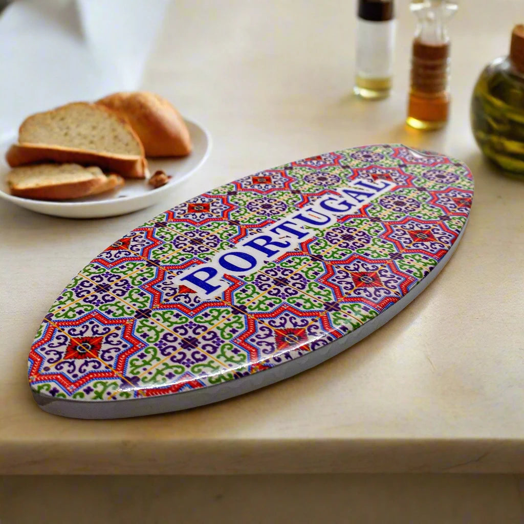 An oval ceramic souvenir decorated with a vibrant mosaic pattern reminiscent of Portuguese azulejo tiles. The word "PORTUGAL" is centered in bold blue uppercase letters. The item has a cork base, casting a light shadow on a white background.