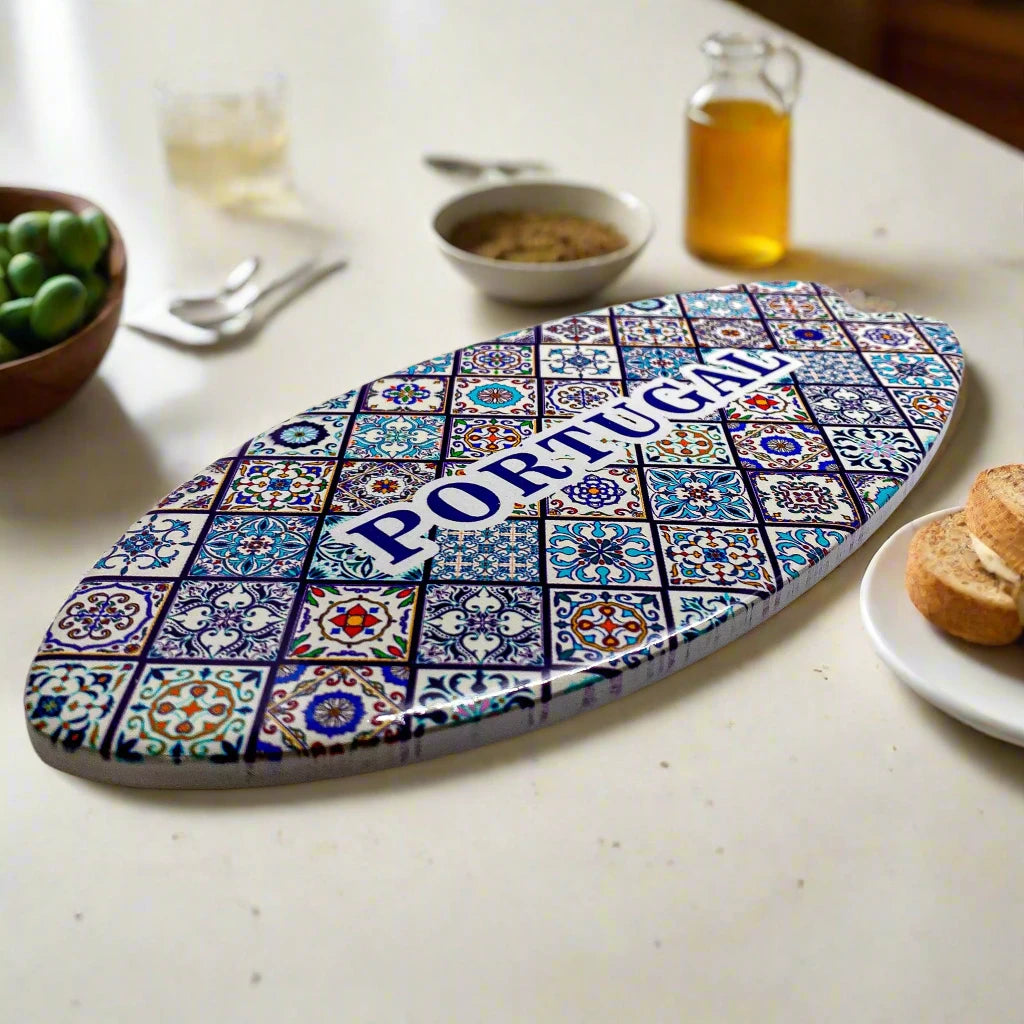 An oval ceramic piece with a mosaic pattern showcasing traditional Portuguese azulejo tiles. "PORTUGAL" is boldly displayed in blue uppercase letters at the center. The souvenir is set on a cork base and casts a faint shadow on the white background.