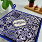 A square ceramic tile showcasing a detailed blue and white pattern, with the word "Portugal" prominently displayed at the center. The tile is bordered by a blue braided cord, has a cork base for durability, and includes a loop at the top for easy hanging.