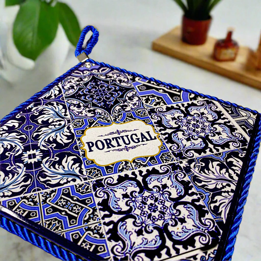 A square ceramic tile showcasing a detailed blue and white pattern, with the word "Portugal" prominently displayed at the center. The tile is bordered by a blue braided cord, has a cork base for durability, and includes a loop at the top for easy hanging.