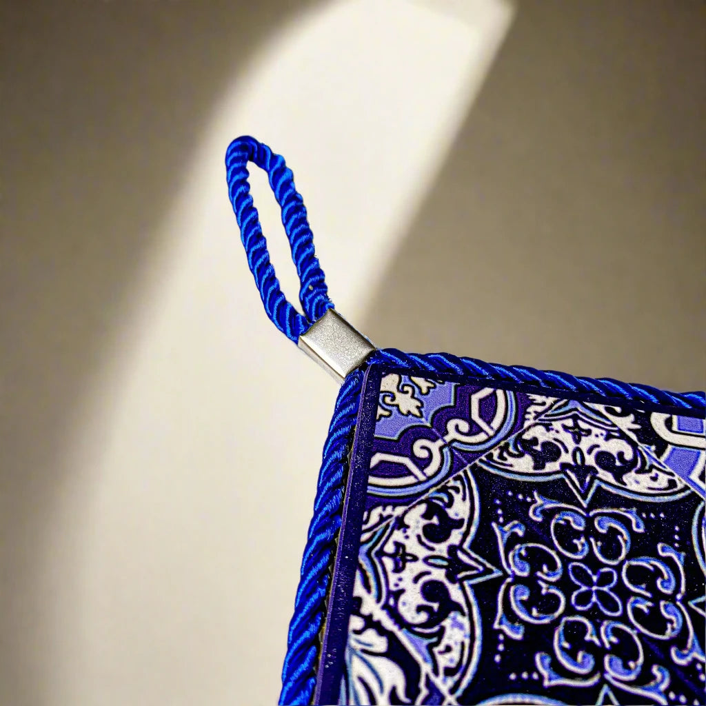 A ceramic tile in a square shape, decorated with a delicate blue and white pattern. The word "Portugal" is centered, framed by a blue braided cord. The tile is supported by a cork base and includes a hanging loop at the top.