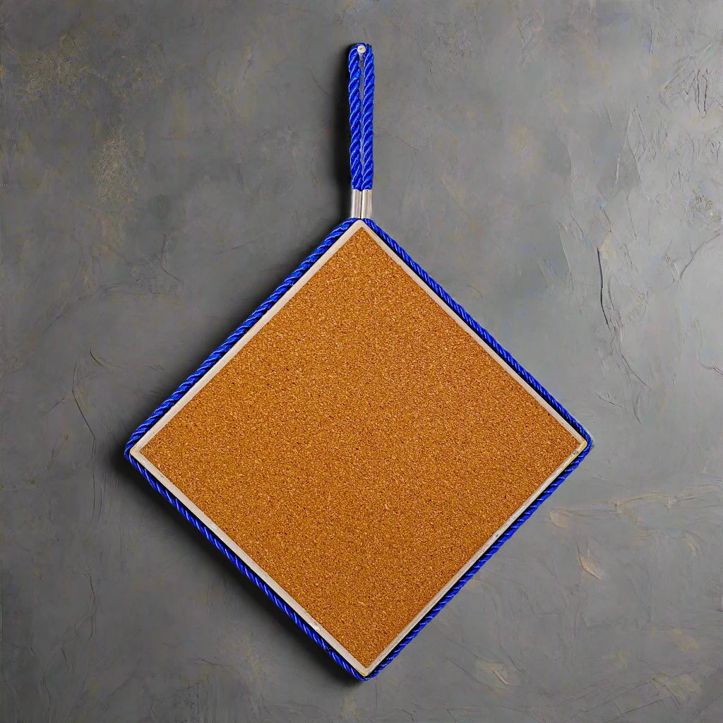 A square cork board mounted on a wall with the word "PORTUGAL" printed in black letters in the bottom right corner, serving as a charming souvenir. Subtle shadows fall across the wall and board, suggesting natural light from a nearby window.