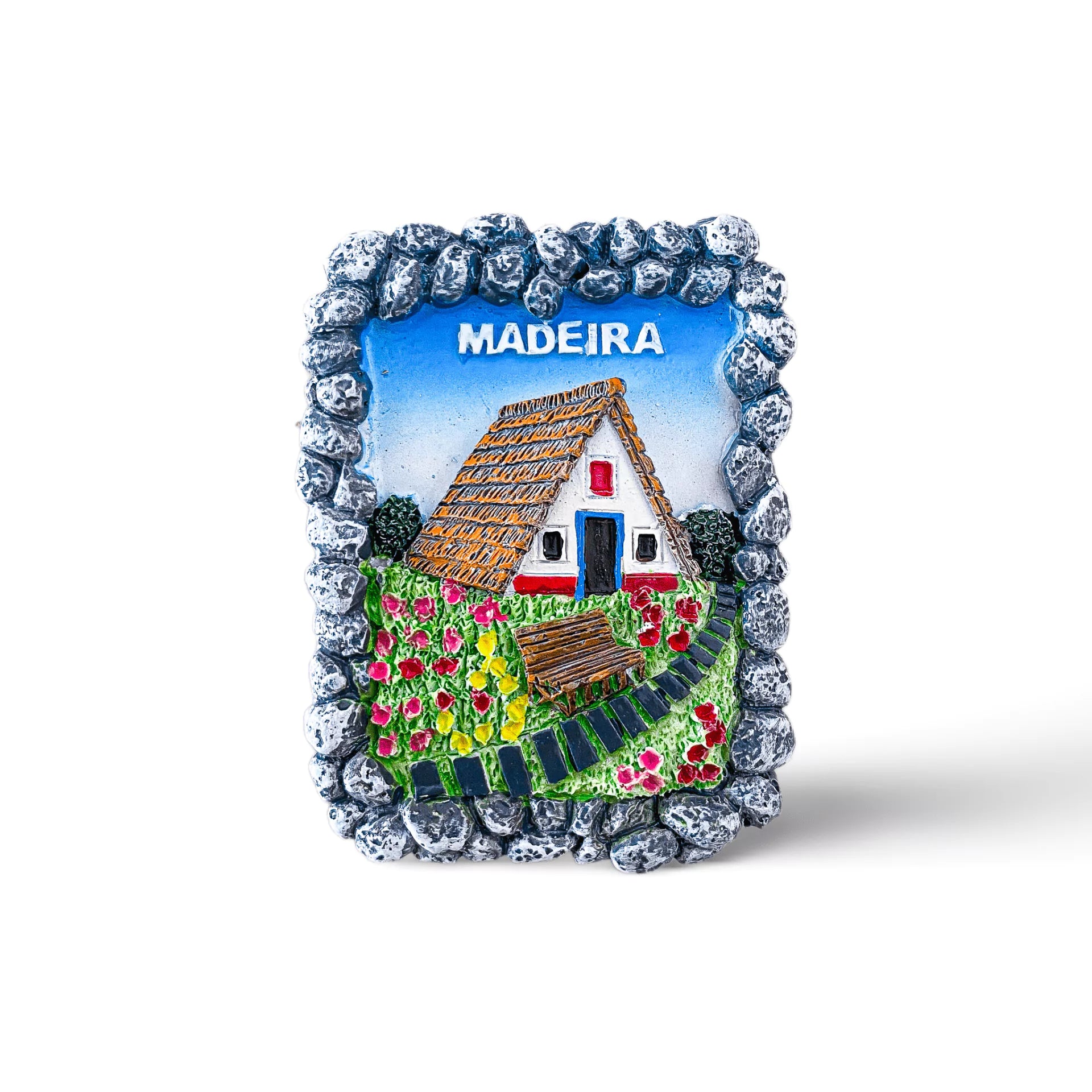 A colorful magnet depicting a traditional house from Madeira, Portugal, with a thatched roof. The magnet is bordered by a stone-like frame and features vibrant flowers and greenery in the foreground. The word "MADEIRA" is written at the top, making it a charming souvenir.