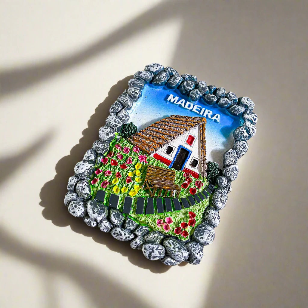 A vibrant magnet showcasing a traditional Madeira house with its iconic thatched roof. The scene is framed by a stone-like border, with bright flowers and lush greenery in the foreground. The word "MADEIRA" is displayed at the top, making this a charming and picturesque souvenir from the Portuguese island.