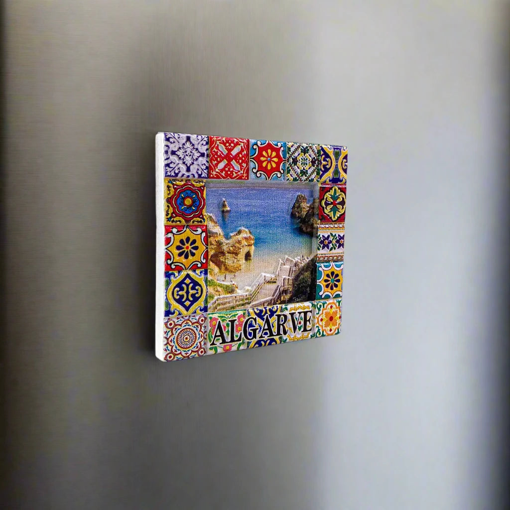 A square picture frame adorned with vibrant, detailed tile designs encases a photo of a coastal beach scene featuring cliffs and clear blue water. The word "Algarve" is inscribed at the bottom. This striking magnet perfectly captures the beauty of the Algarve coastline.