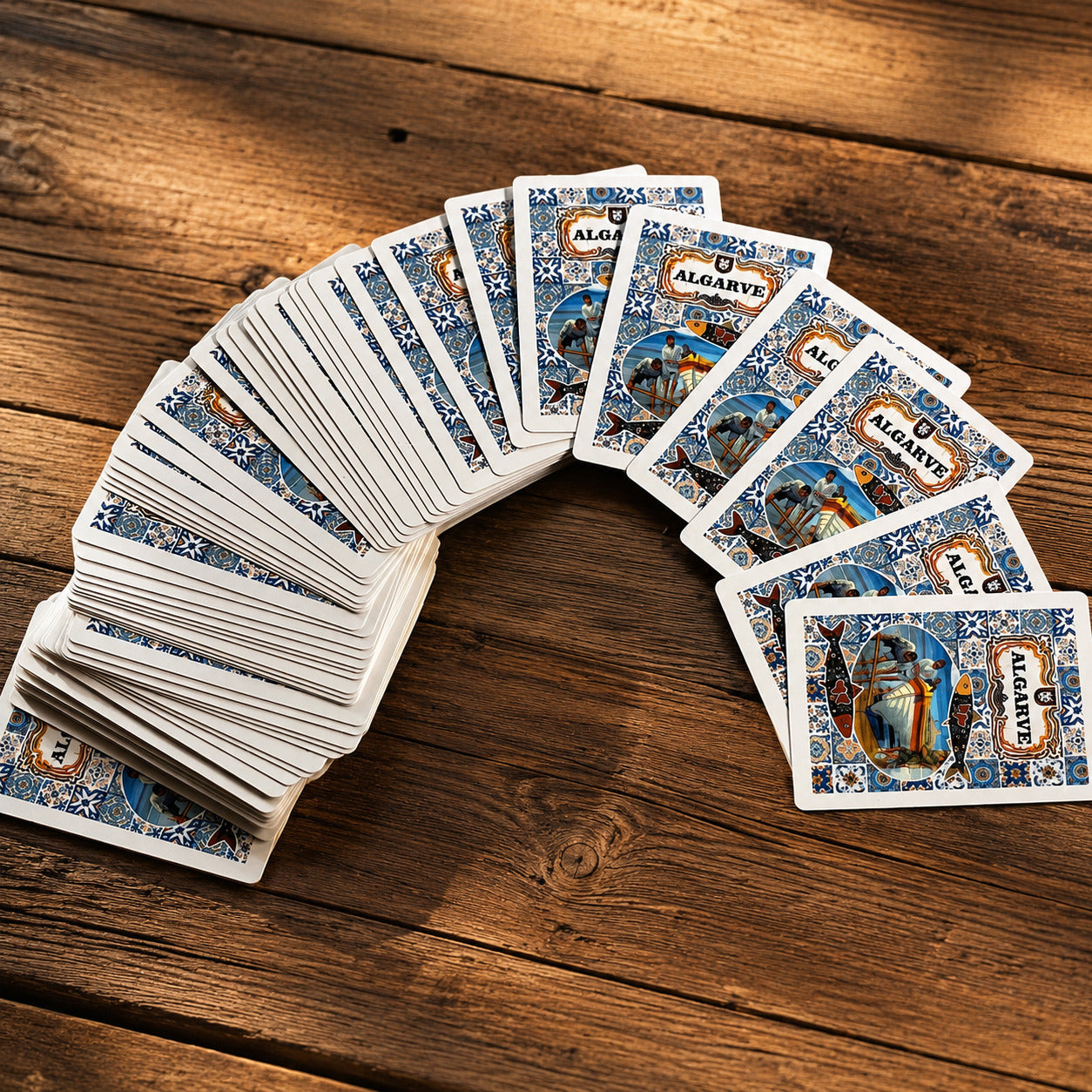 Algarve souvenir playing cards spread on a rustic wooden table, showing an azulejo-inspired back design with sardines and traditional Portuguese fishing artwork.