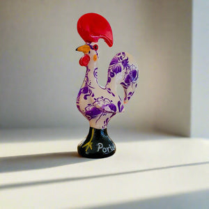 A small ceramic rooster souvenir featuring a purple and white design, symbolizing the Galo de Barcelos from Portugal.