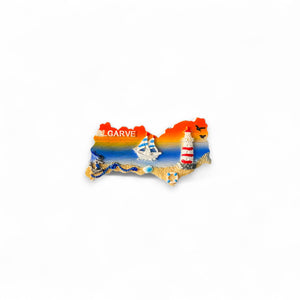 A colorful fridge magnet in the shape of the Algarve region, featuring nautical elements such as a sailboat, lighthouse, and seashells against a sunset or sunrise background.