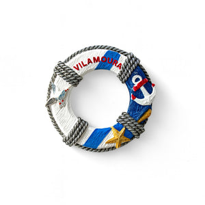 A round Vilamoura souvenir magnet featuring a classic lifebuoy design with nautical symbols such as a rope, starfish, and anchor, and the name 'Vilamoura' written in red.