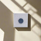 A small square canvas with a textured, off-white surface and a dark circular shape in the center is mounted on a beige wall. Shadows from an unseen magnet cast diagonal lines across the canvas and wall, creating a geometric pattern reminiscent of an abstract souvenir.