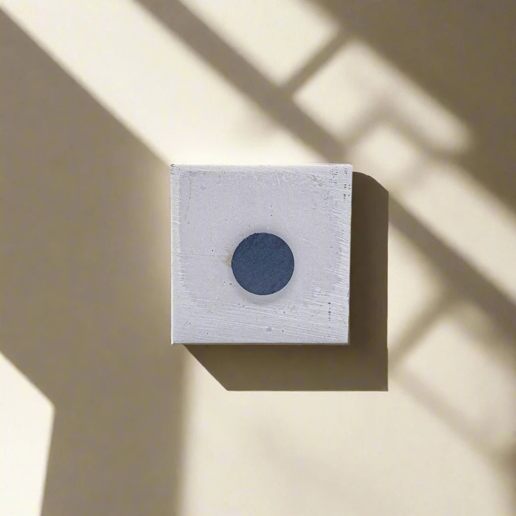 A small square canvas with a textured, off-white surface and a dark circular shape in the center is mounted on a beige wall. Shadows from an unseen magnet cast diagonal lines across the canvas and wall, creating a geometric pattern reminiscent of an abstract souvenir.