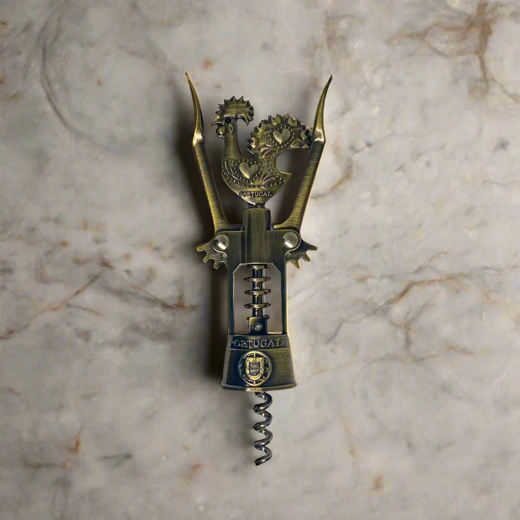 A bronze corkscrew is shown, intricately designed with a handle that resembles the Barcelos Rooster, a symbol synonymous with Portugal. The rooster's handle has a heart motif, and "Portugal" is engraved on the midsection. This souvenir includes two levers and a turning mechanism.